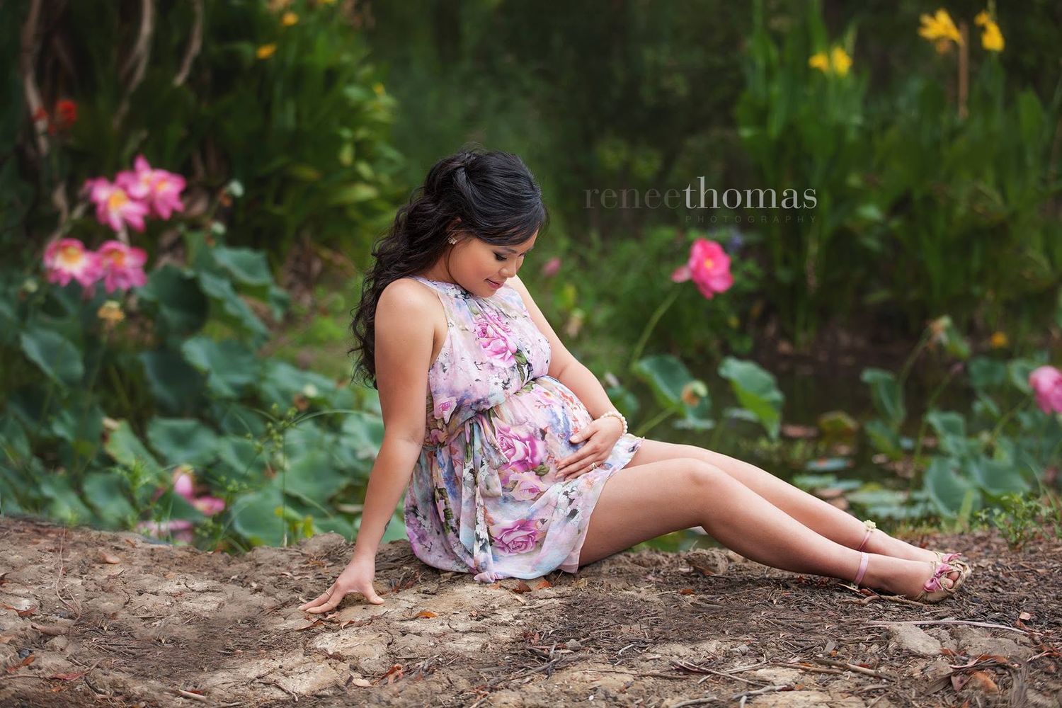 Pregnant woman in floral dress sitting in a garden surrounded by colorful flowers.