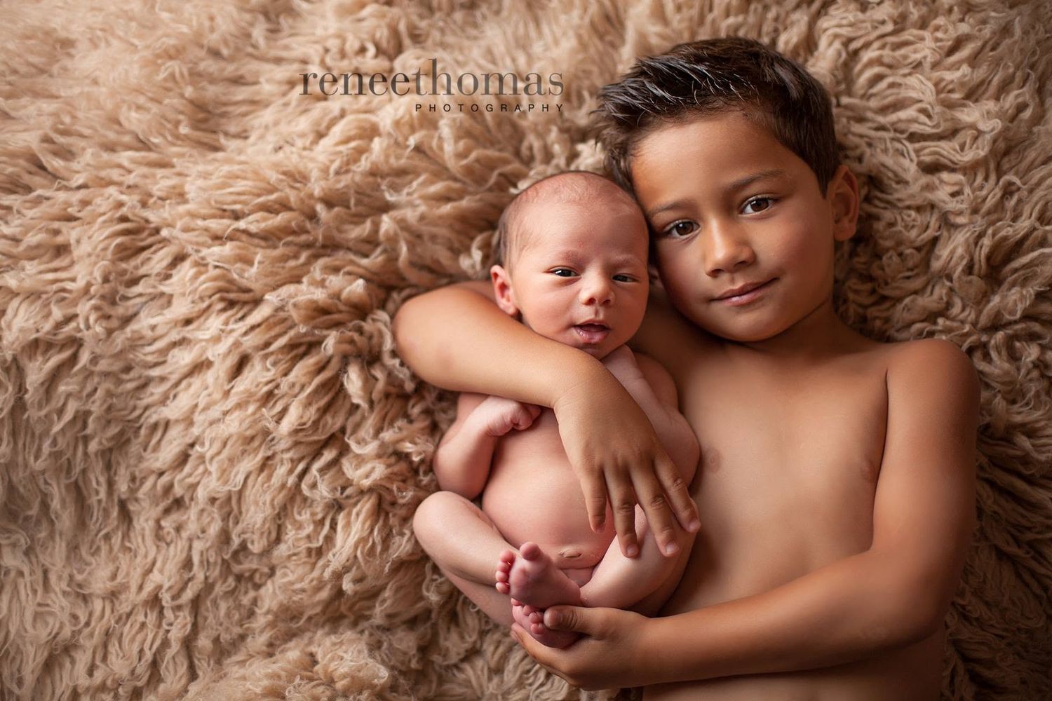 Young boy lying on fluffy beige fur rug gently holding a sleeping newborn sibling.