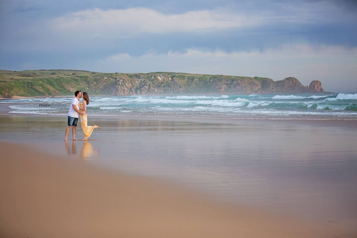 Couple standing together on a sandy beach with dramatic ocean waves and rocky cliffs in the background.