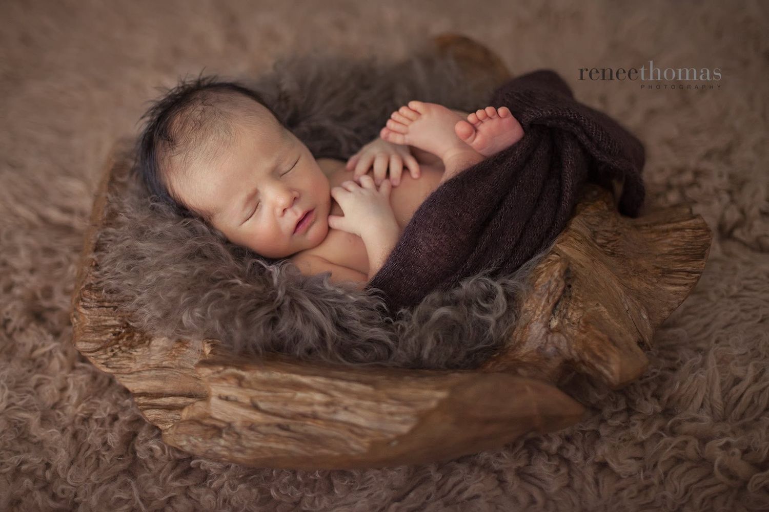 Sleeping newborn baby curled in wooden bowl with gray fur, wrapped in brown knit wrap.