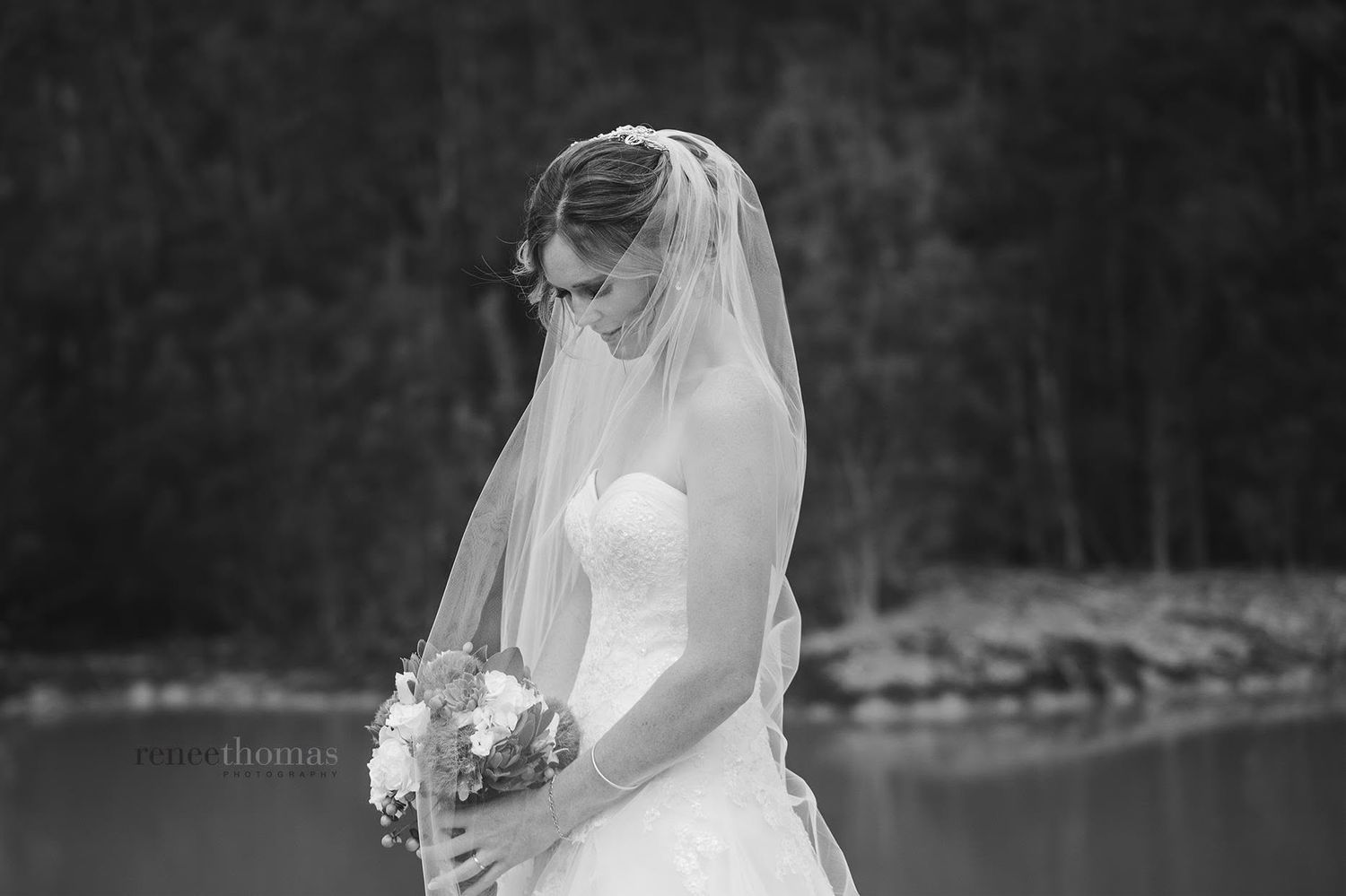 Black and white portrait of a bride in a strapless gown and veil holding a bouquet by a lake.