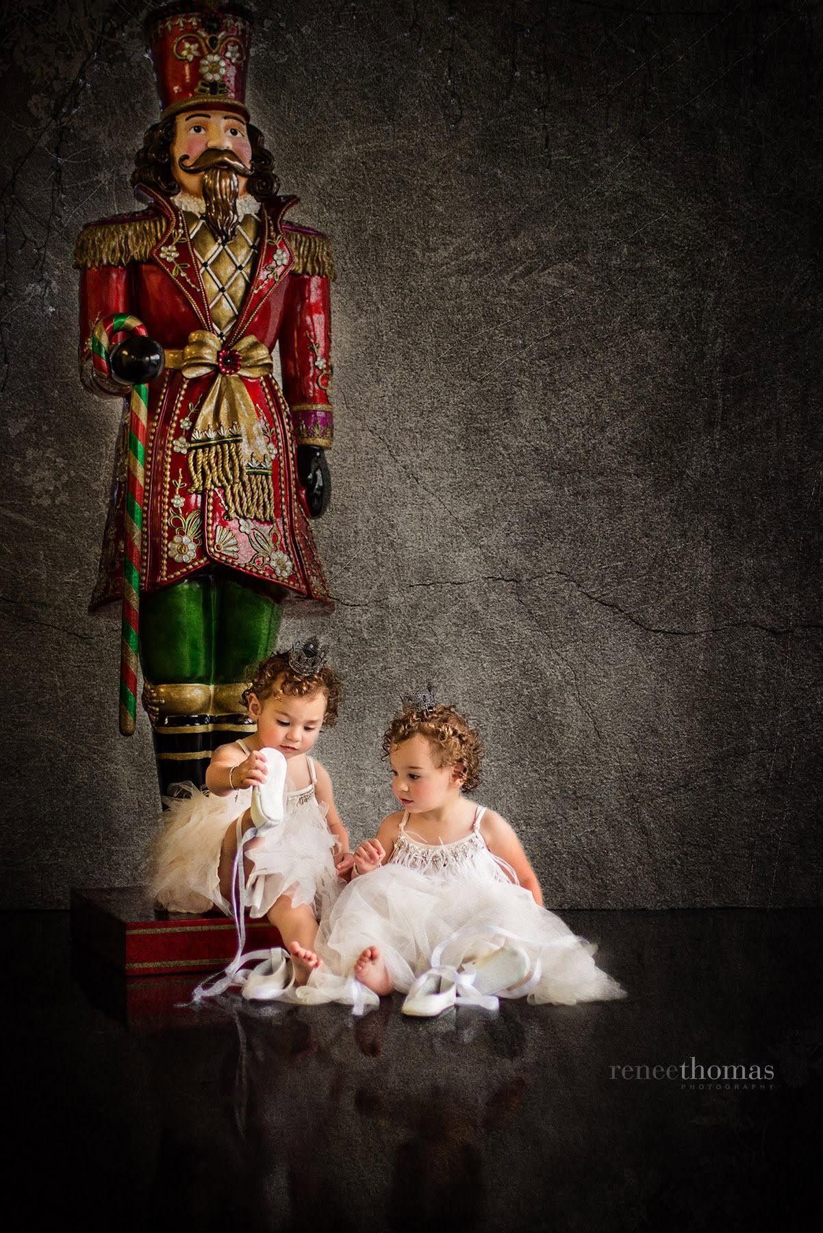 Two toddler girls in white tutus sitting beside a large nutcracker holiday decoration by Renee Thomas.