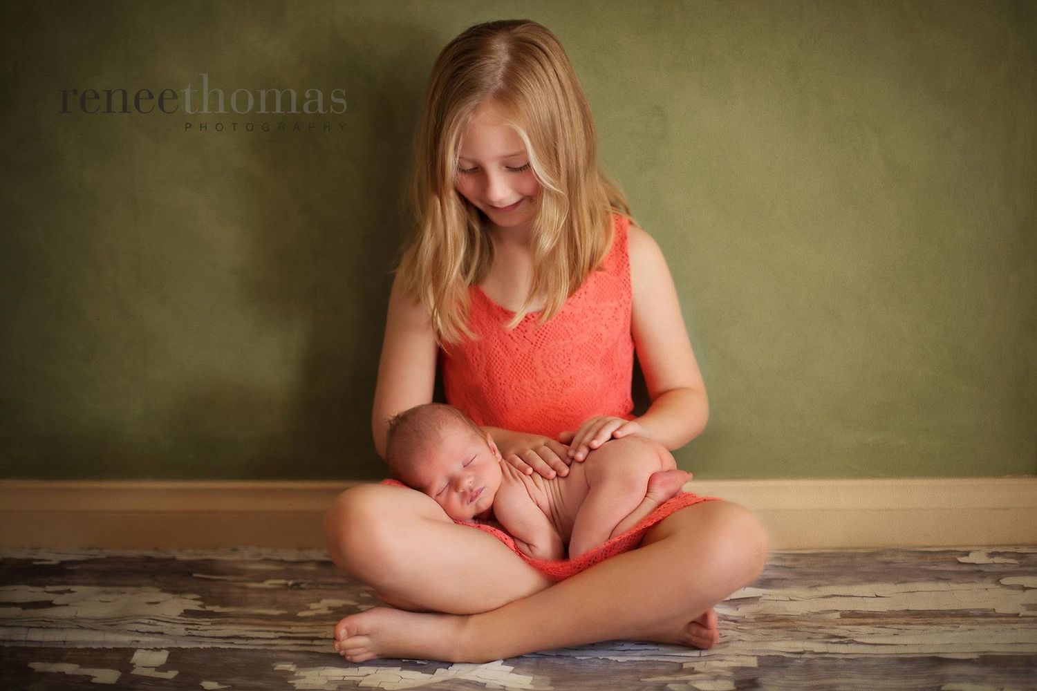 Young girl in orange dress sitting cross-legged holding a sleeping newborn baby.