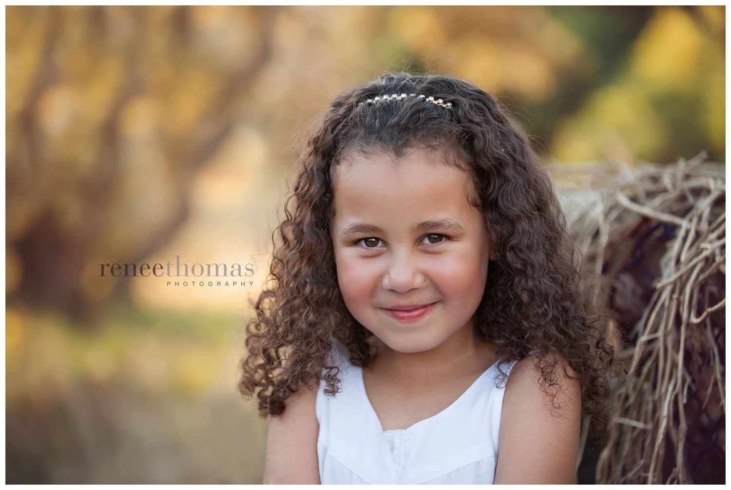Young girl with curly hair wearing a white dress and headband smiling outdoors in warm autumn light.