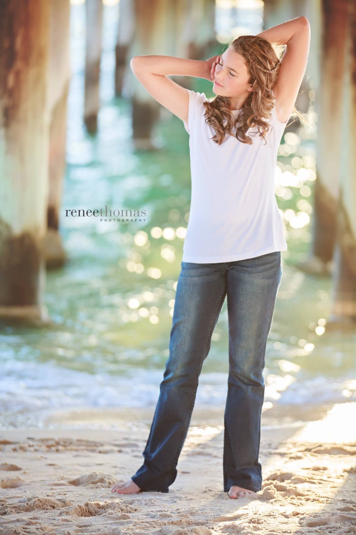 Young woman posing barefoot on sandy beach under pier with turquoise water backdrop.