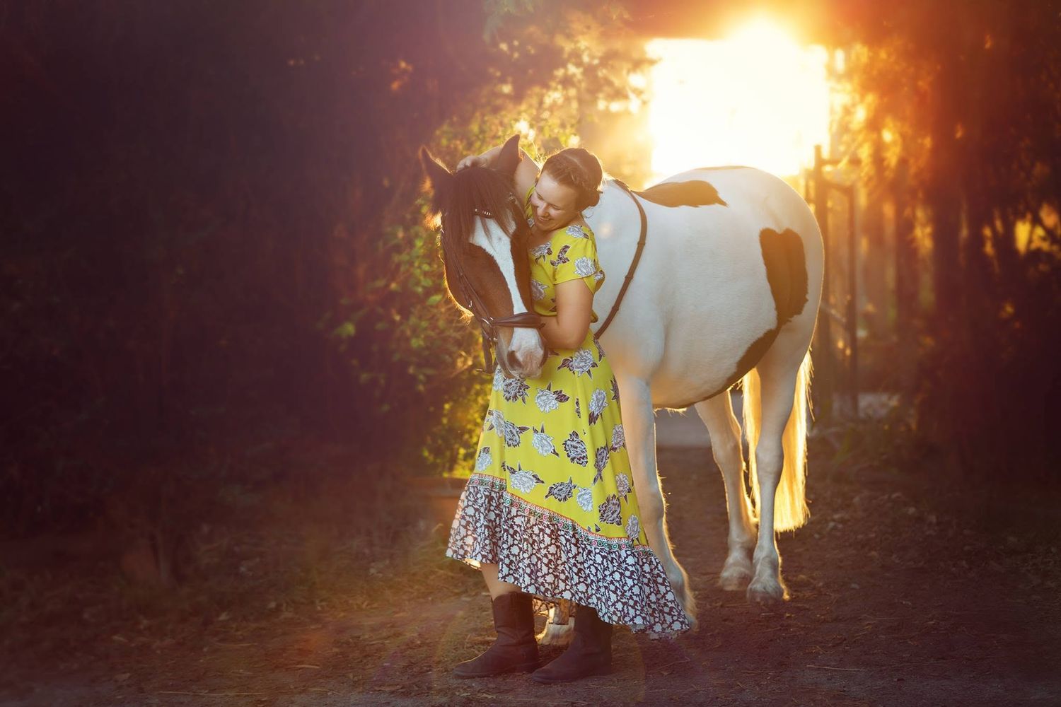 Woman in yellow floral dress kissing a paint horse at golden sunset in a scenic forest path.