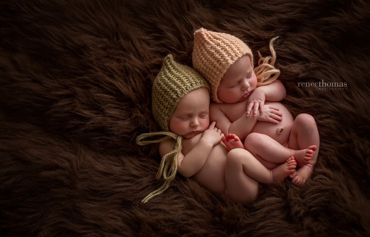 Two sleeping newborn twins wearing knitted bonnets cuddled together on a fluffy brown fur blanket.