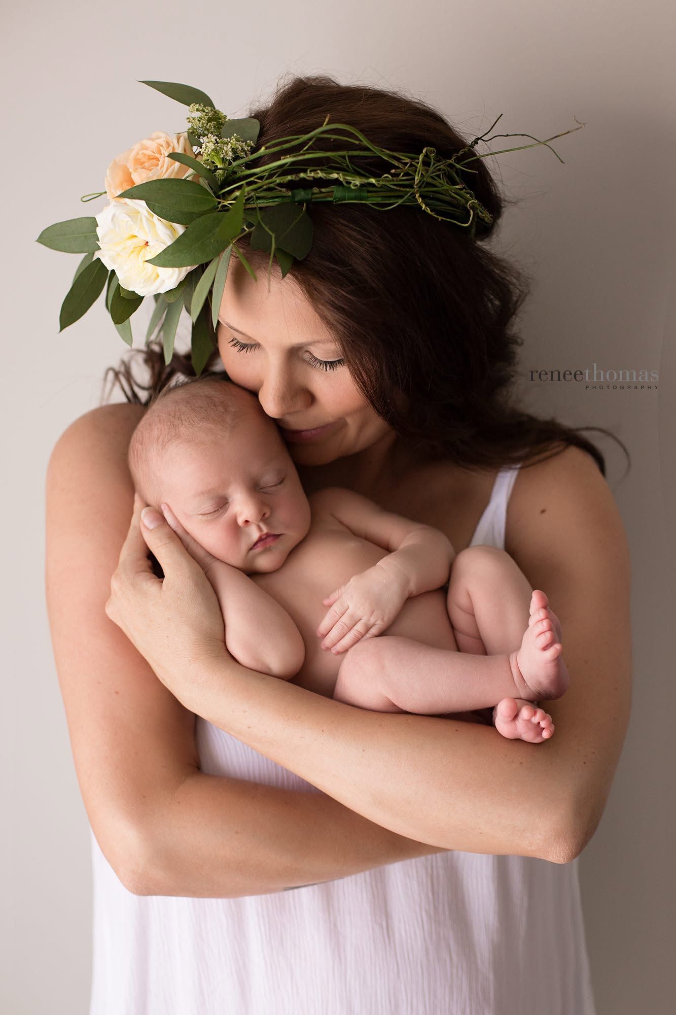 Mother wearing floral crown tenderly holding sleeping newborn baby in a studio portrait.