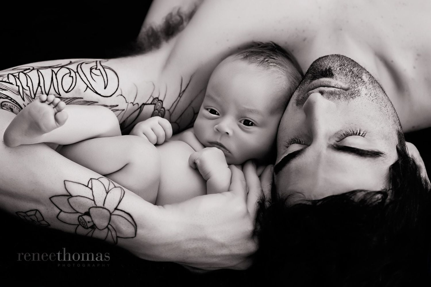 Black and white photo of tattooed parents lying close together with their newborn baby.