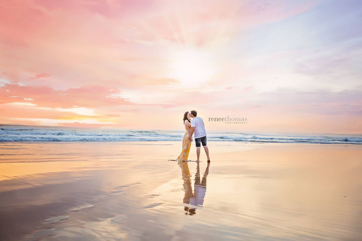 Couple sharing romantic kiss on reflective beach at stunning pink and purple sunset.