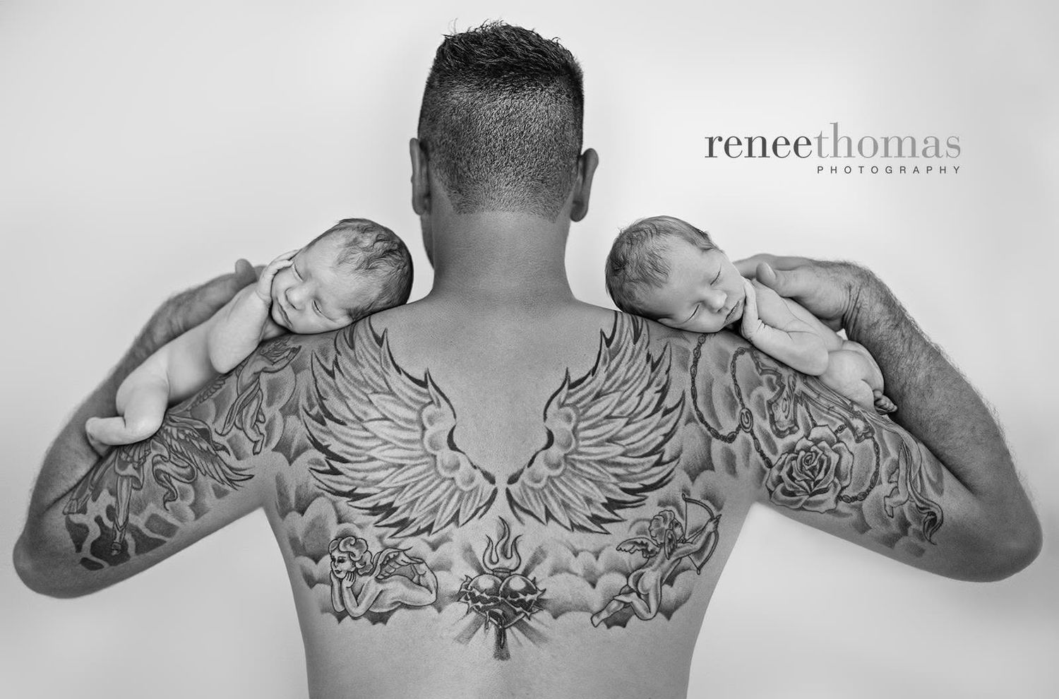 Tattooed father holding twin newborns on his shoulders in black and white portrait.