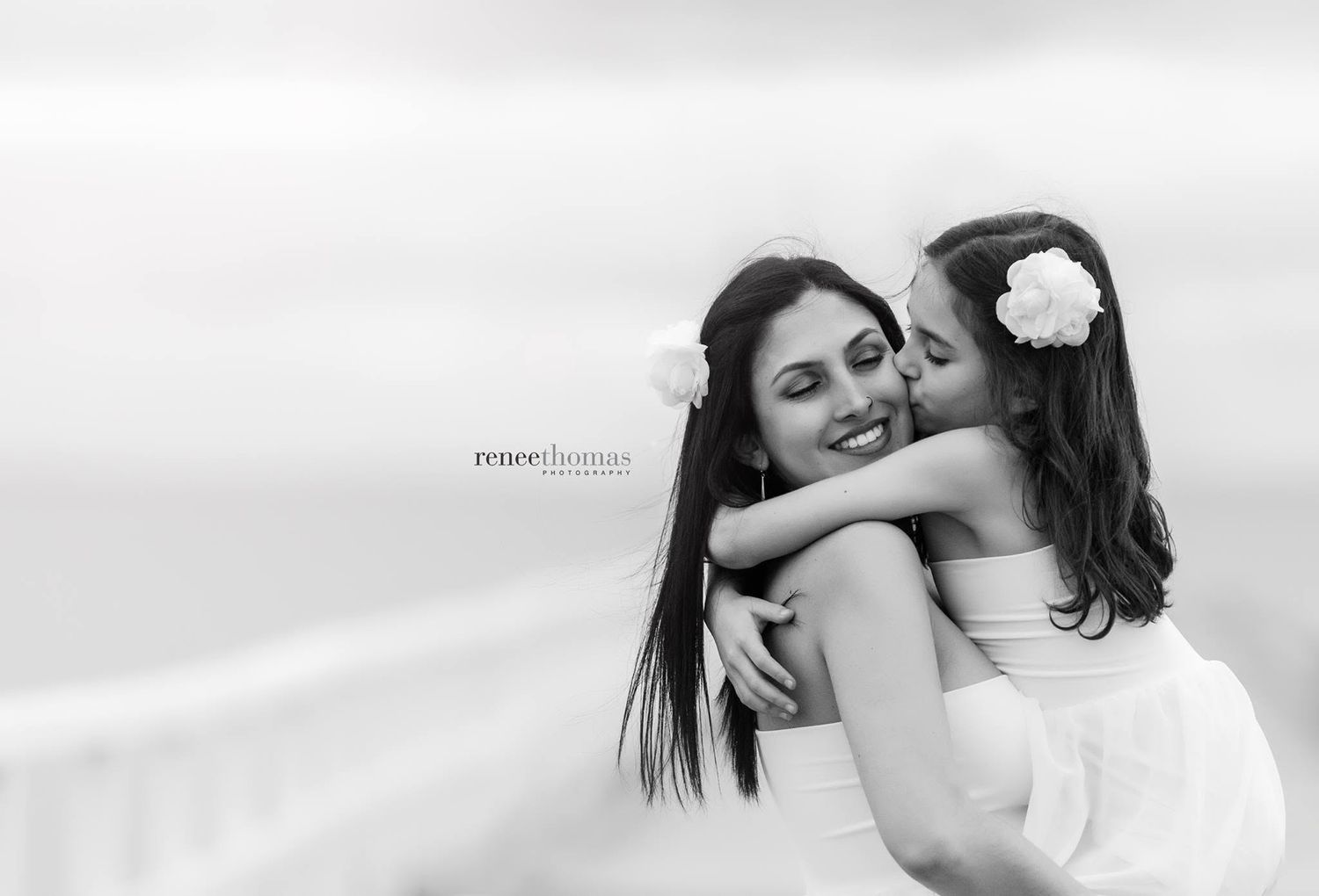 Black and white photo of a smiling woman being kissed on the cheek by a young girl outdoors.