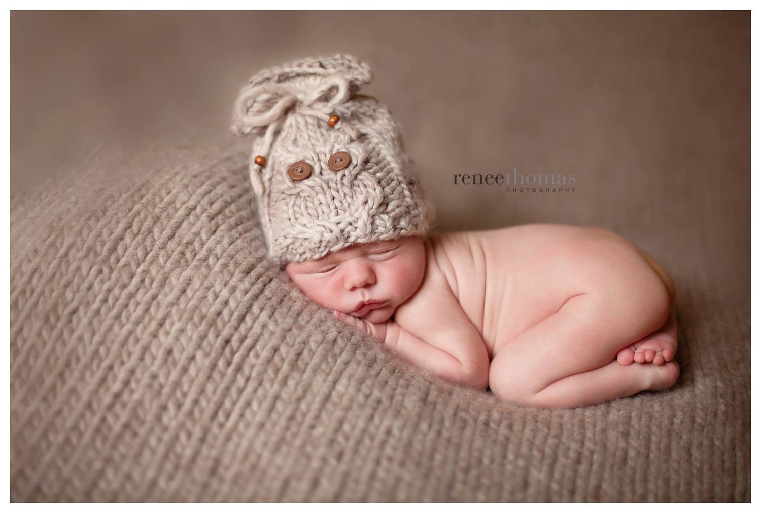 Sleeping newborn baby wearing a cream knitted owl hat on a brown textured blanket.