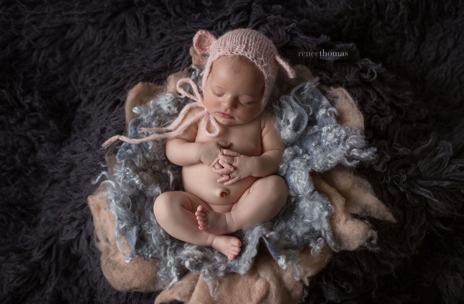 Sleeping newborn baby in pink bear bonnet posed on layered gray and floral textured blankets.