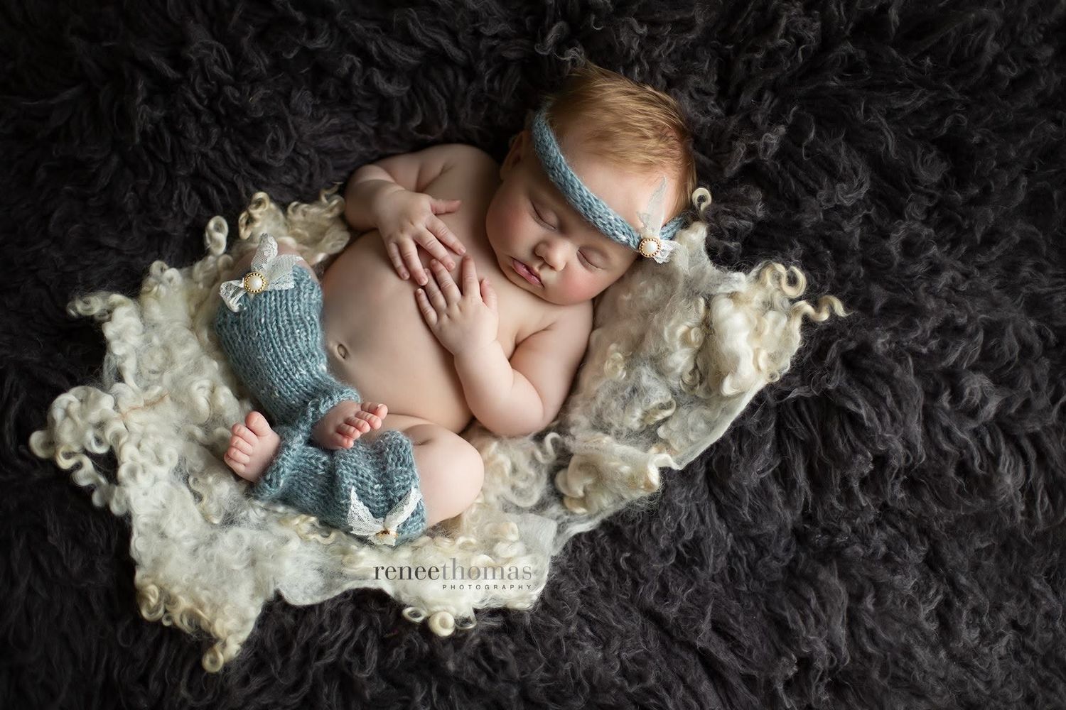 Sleeping red-haired newborn baby in blue knit outfit resting on a white lace blanket on dark fur.