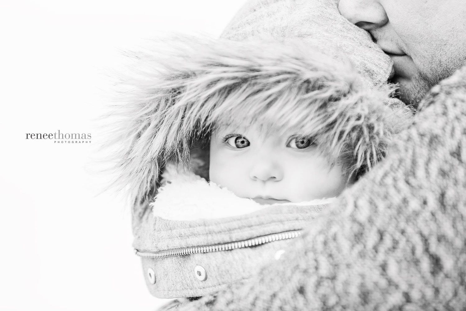 Black and white close-up of a newborn bundled in a fur-trimmed jacket being held by a parent.