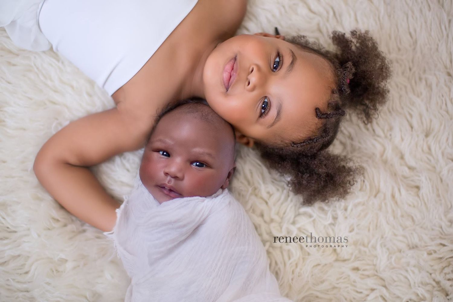Young girl lying beside swaddled newborn sibling on cream fluffy rug, smiling warmly.