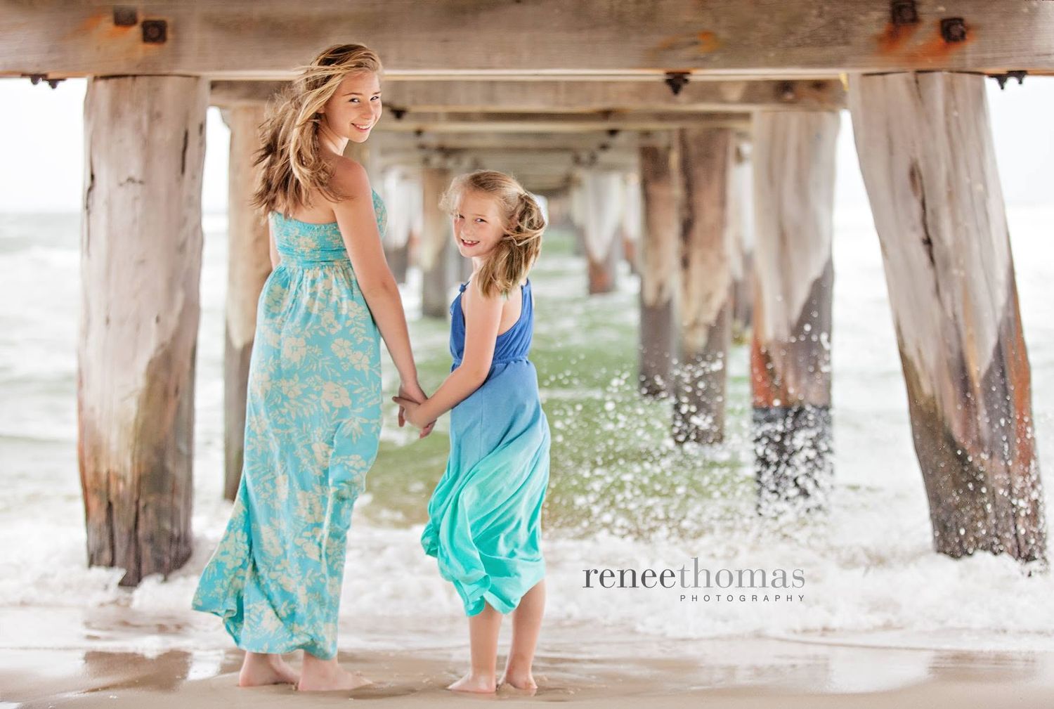 Mother and daughter holding hands and playing in ocean waves under a pier by Renee Thomas Photography.