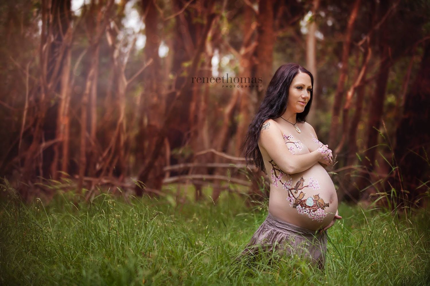 Pregnant woman in sheer dress standing in lush green field surrounded by tall trees.