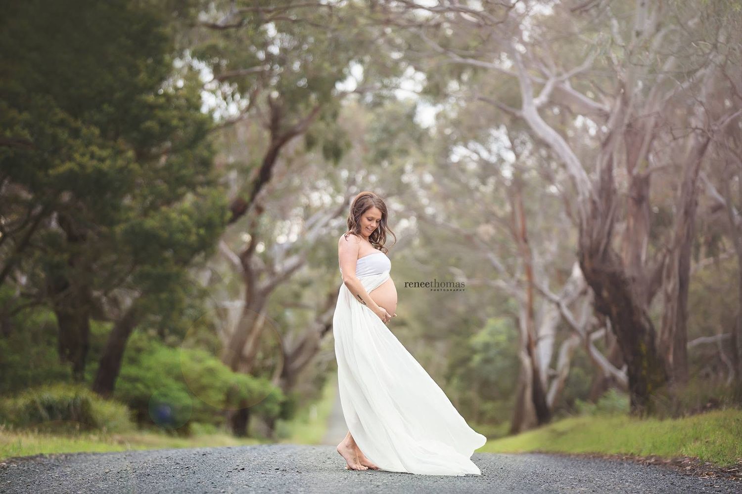 Pregnant woman in white flowing gown standing on a tree-lined road during an outdoor maternity photo session.