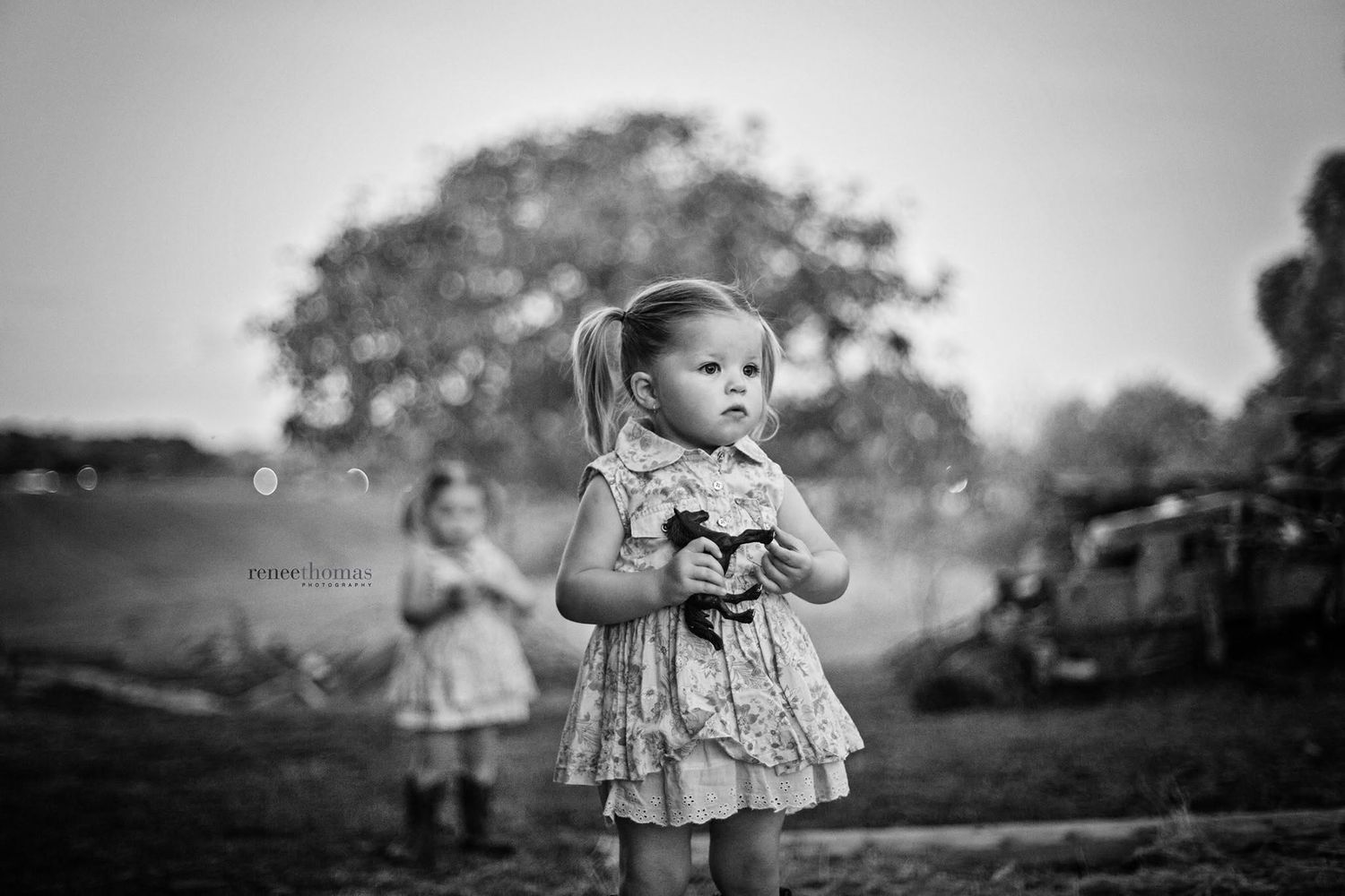 Black and white portrait of a toddler girl in a floral dress holding a toy in an outdoor field.
