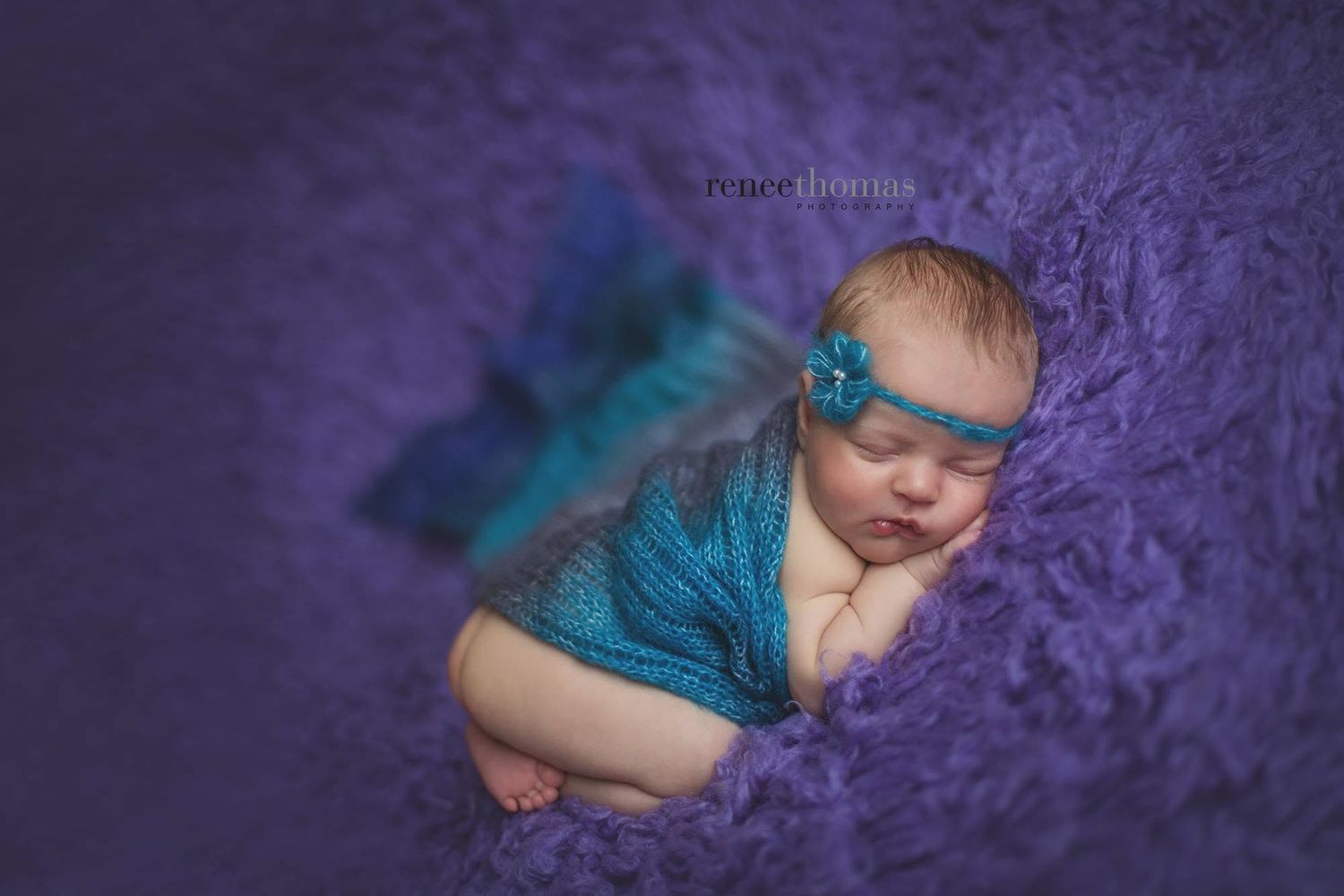 Sleeping newborn baby wrapped in teal knit on purple fluffy blanket with floral headband.