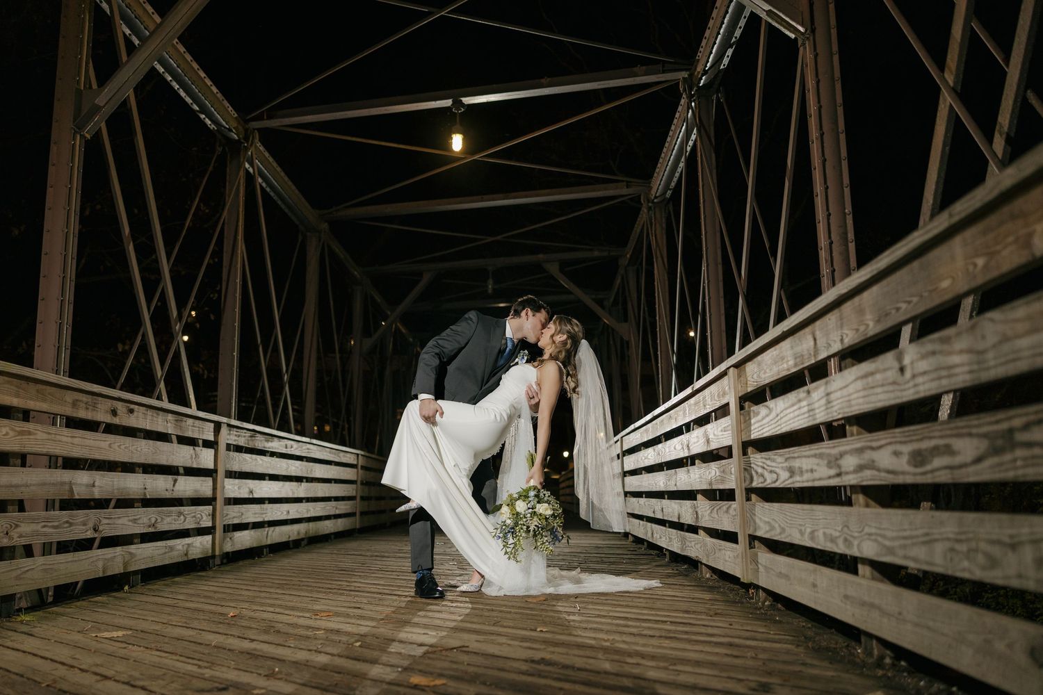 Bride and groom share a romantic dip kiss on a wooden bridge at night during their wedding celebration.