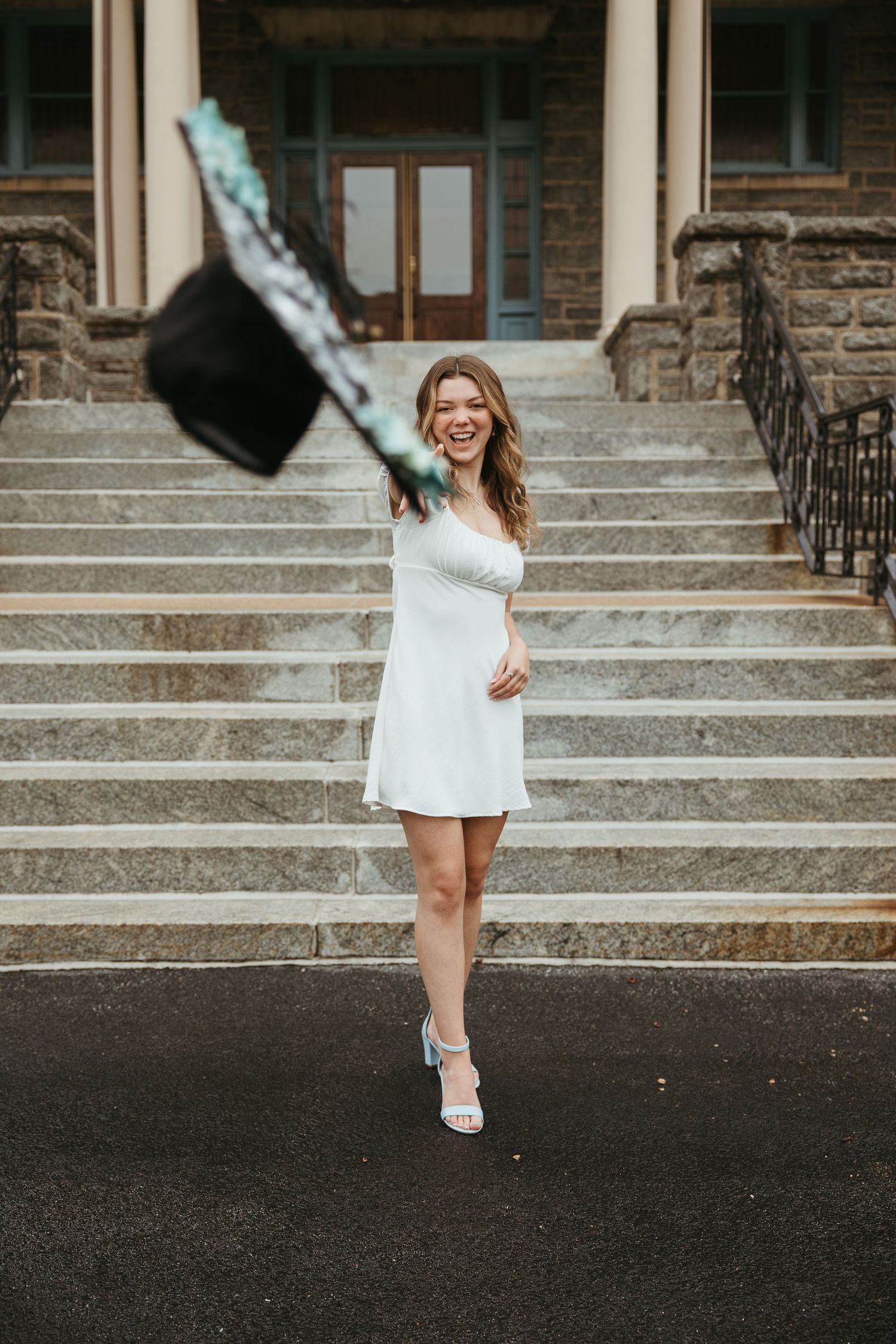Graduate jumps joyfully on stone steps while tossing graduation cap in celebration.