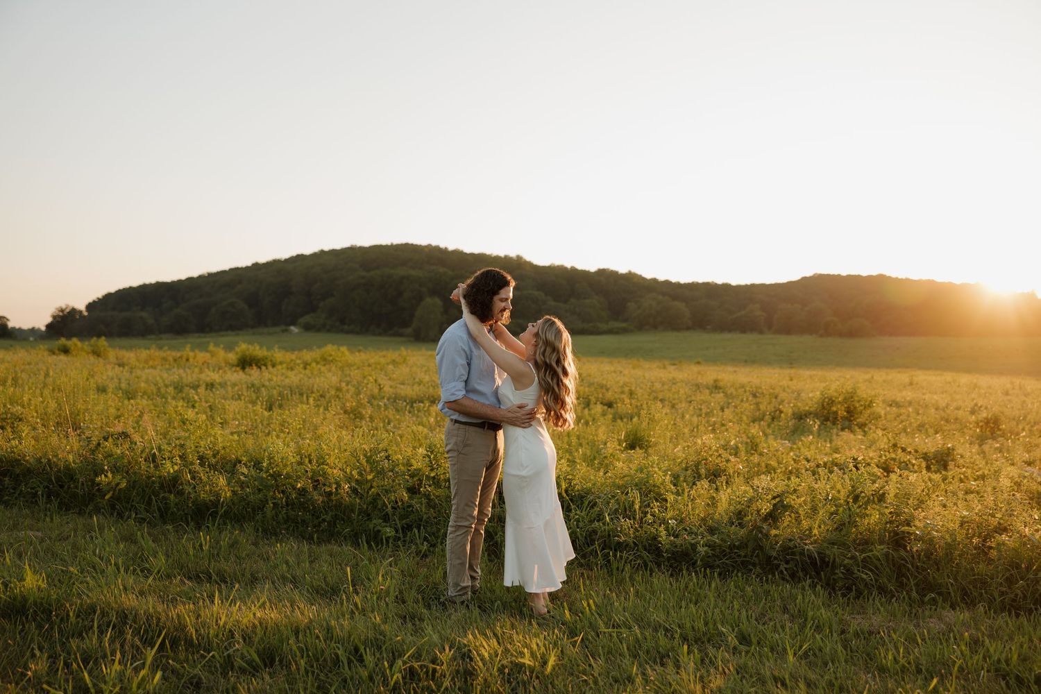A couple embraces in a golden sunlit meadow at sunset with rolling hills in the background.