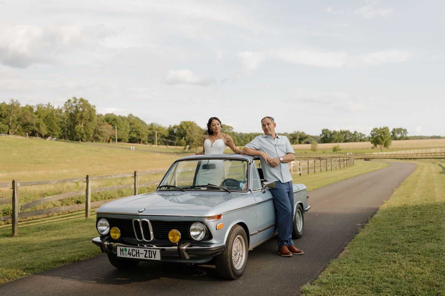 Couple posing with a classic light blue BMW 2002 on a scenic country road surrounded by green fields.