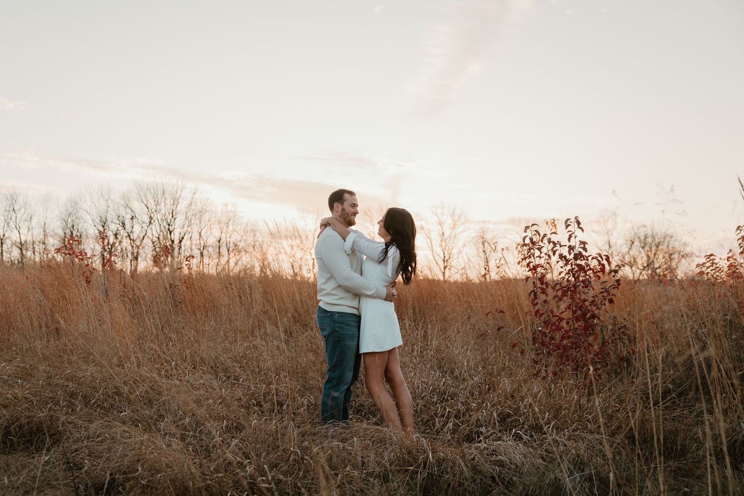 A romantic couple embraces in a field of tall grass during golden hour sunset.