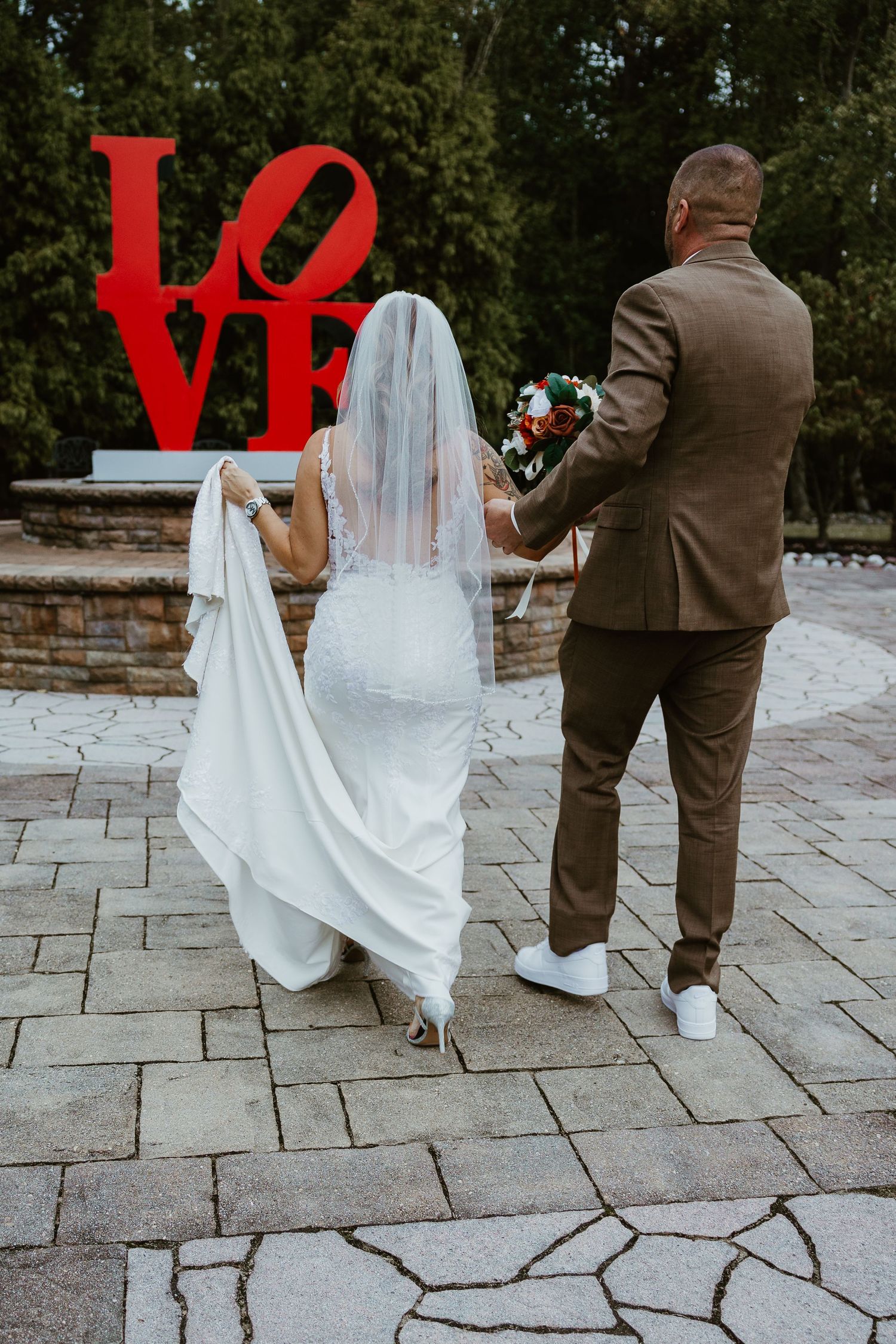 Couple stands before red LOVE sculpture on stone patio during wedding photoshoot.