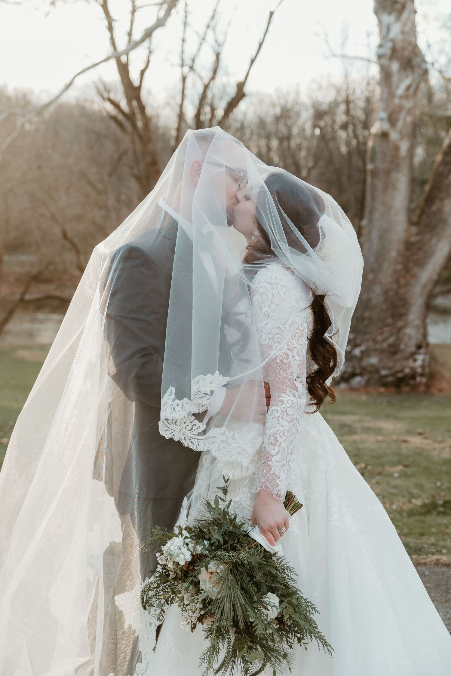 A romantic wedding photo showing the couple embracing under a flowing veil in a natural outdoor setting.