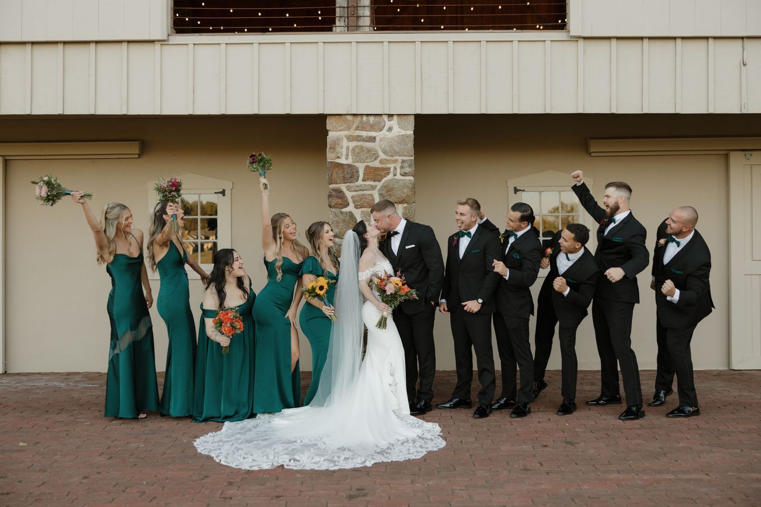 Wedding party in formal attire poses together in front of a light colored building wall.