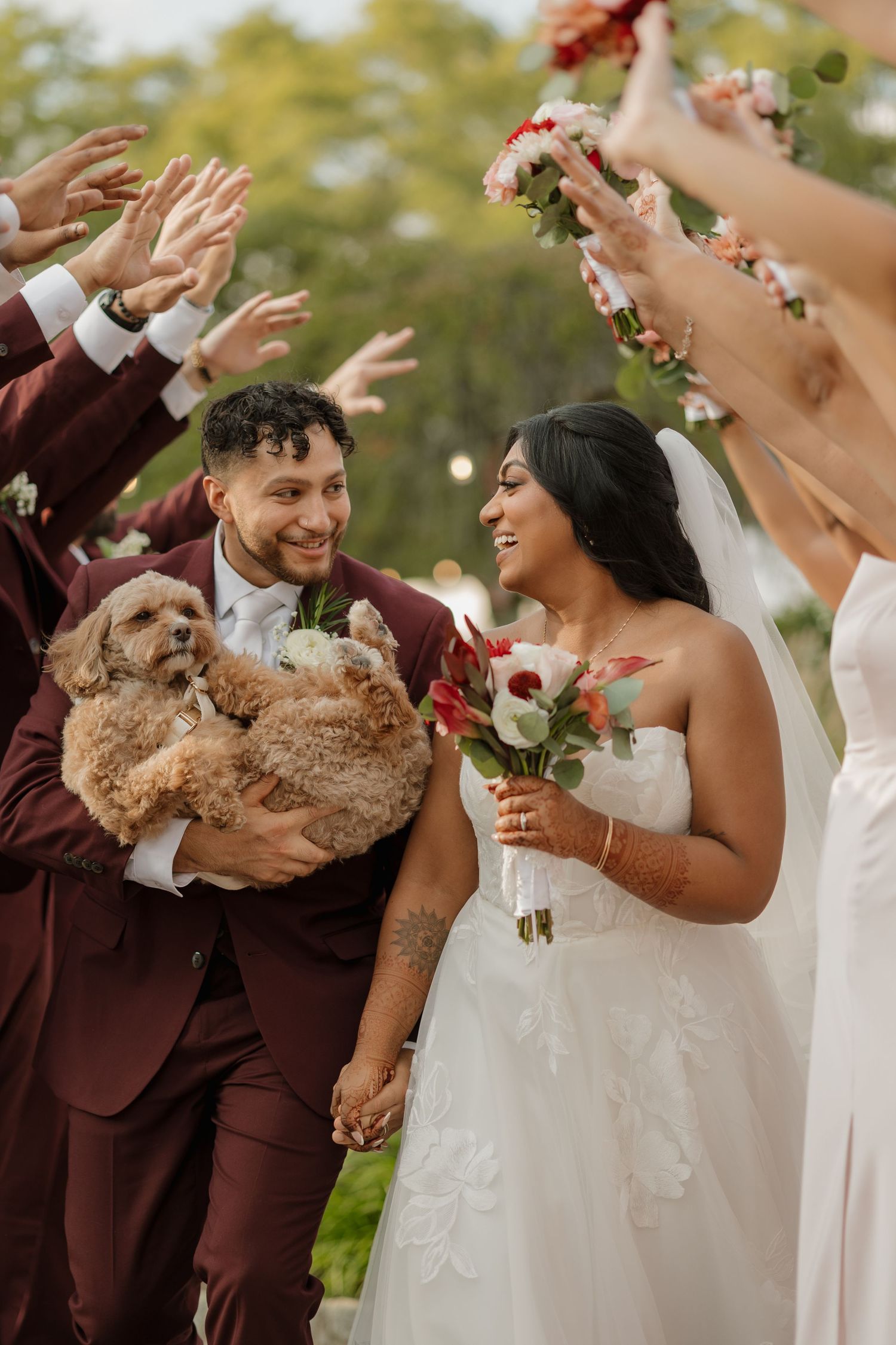 Bride and groom walk through floral arch, groom holds fluffy dog, bride carries red and white bouquet in wedding celebration.