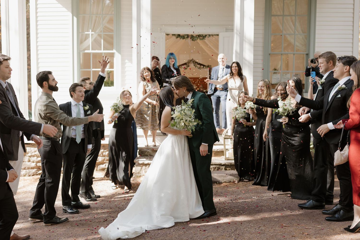 Bride and groom share a kiss during wedding recessional while guests celebrate with confetti in elegant venue.