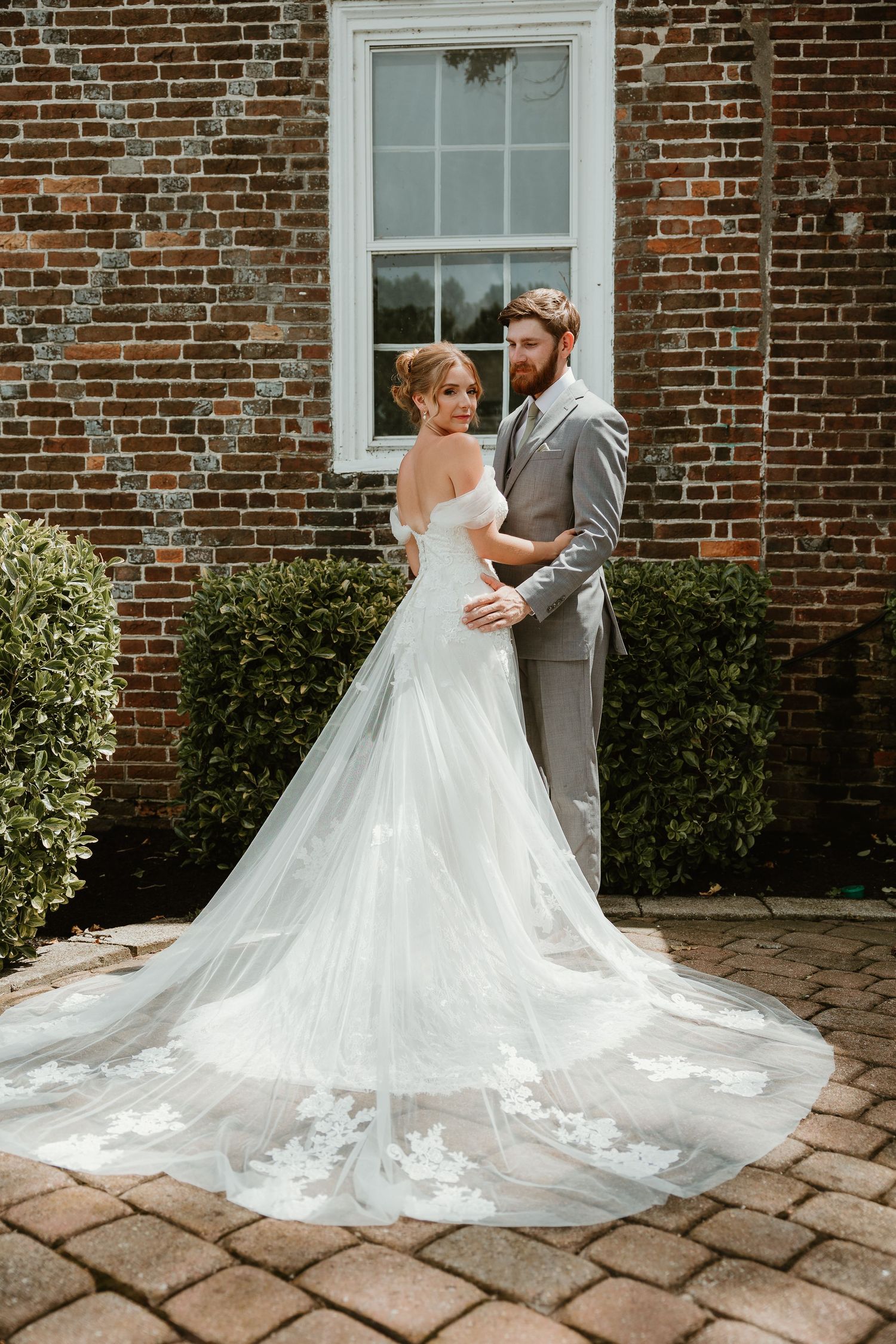 Classic wedding portrait against brick wall showcasing bride's long train and elegant white gown.