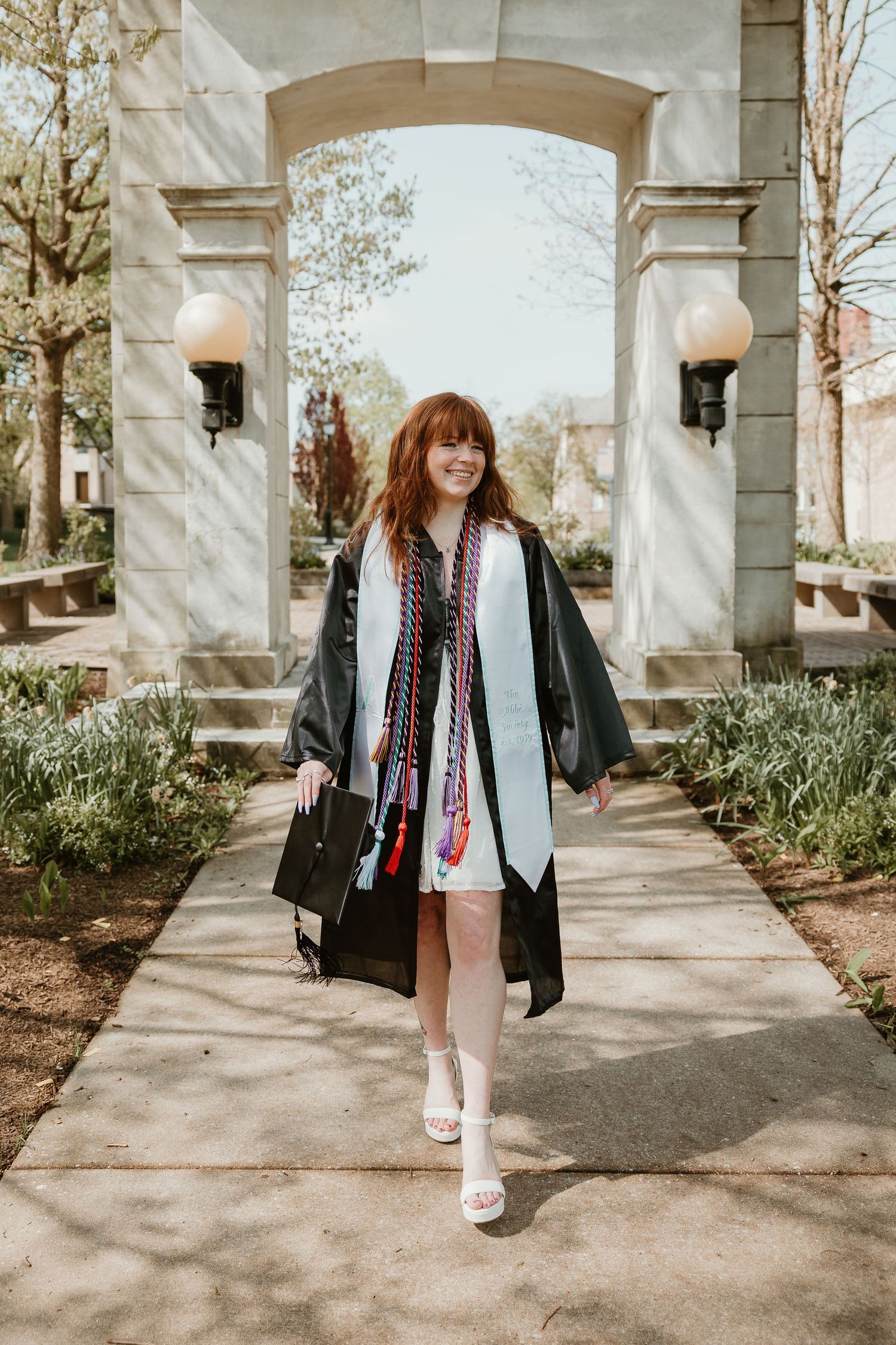 Graduate in a black cap and gown poses joyfully under a stone archway with decorative lanterns.