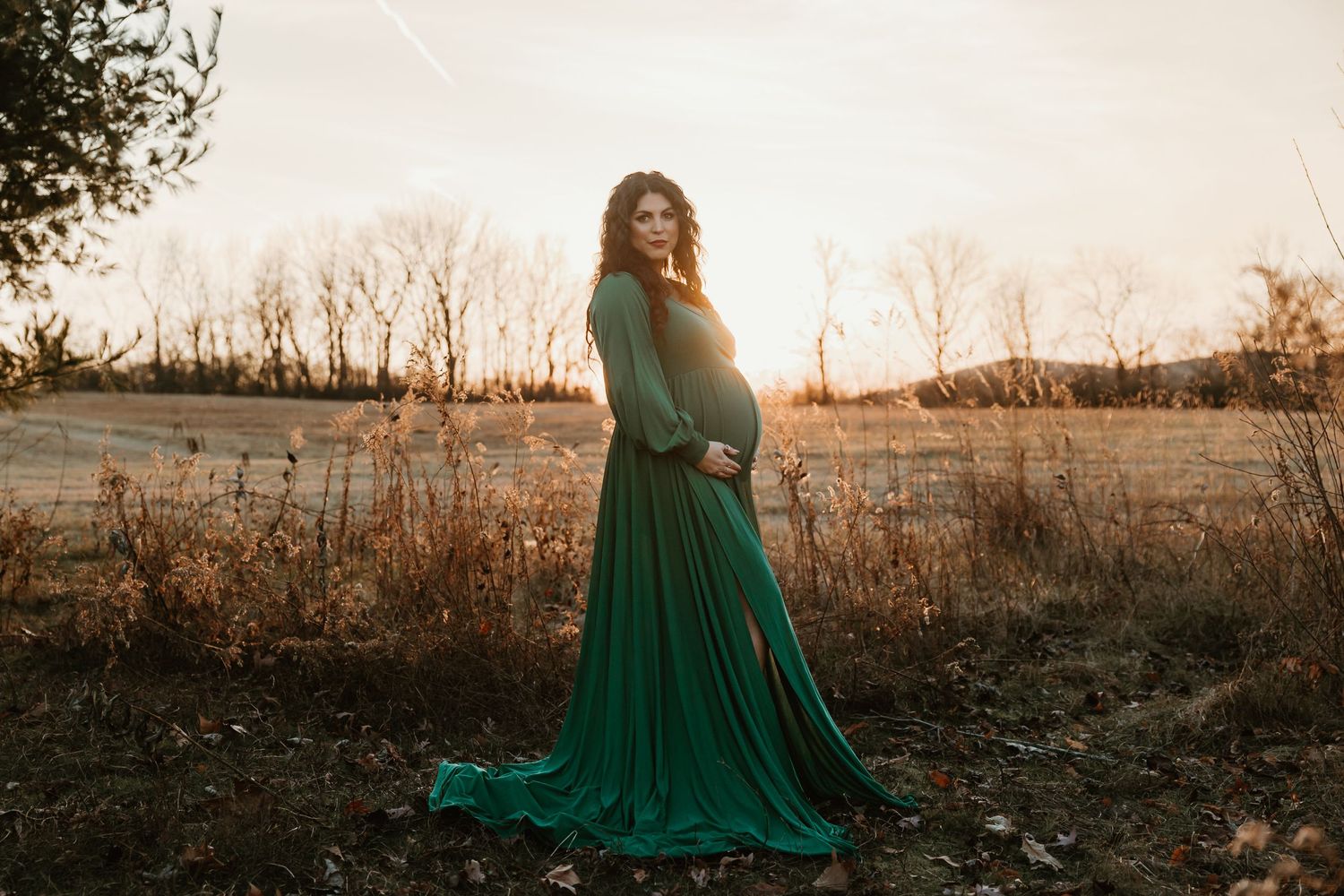 A flowing emerald green maternity gown billows in a field at sunset during a serene outdoor photo session.