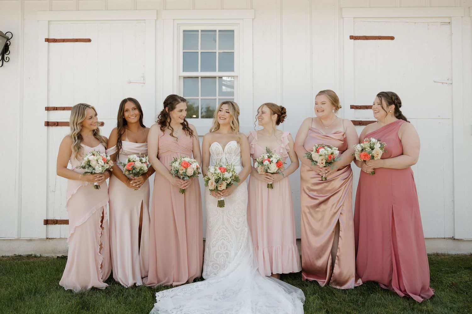 Bride in white lace gown stands with six bridesmaids in blush pink dresses holding bouquets outside a white barn.