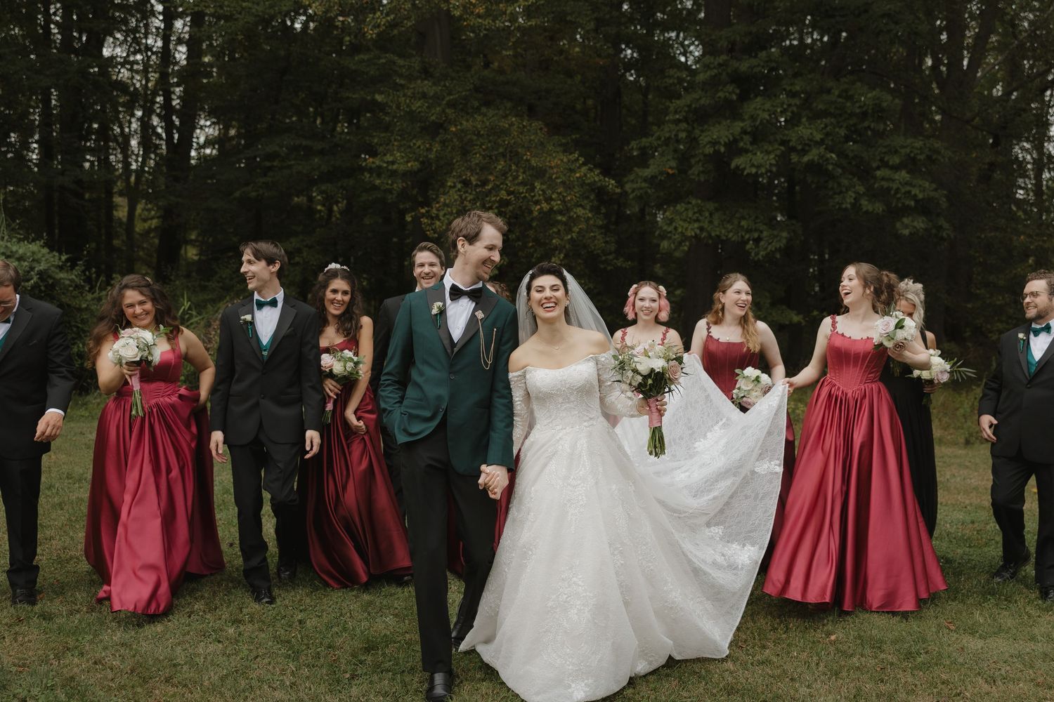 Wedding party in formal attire poses outdoors with bridesmaids in burgundy dresses and groomsmen in dark suits.