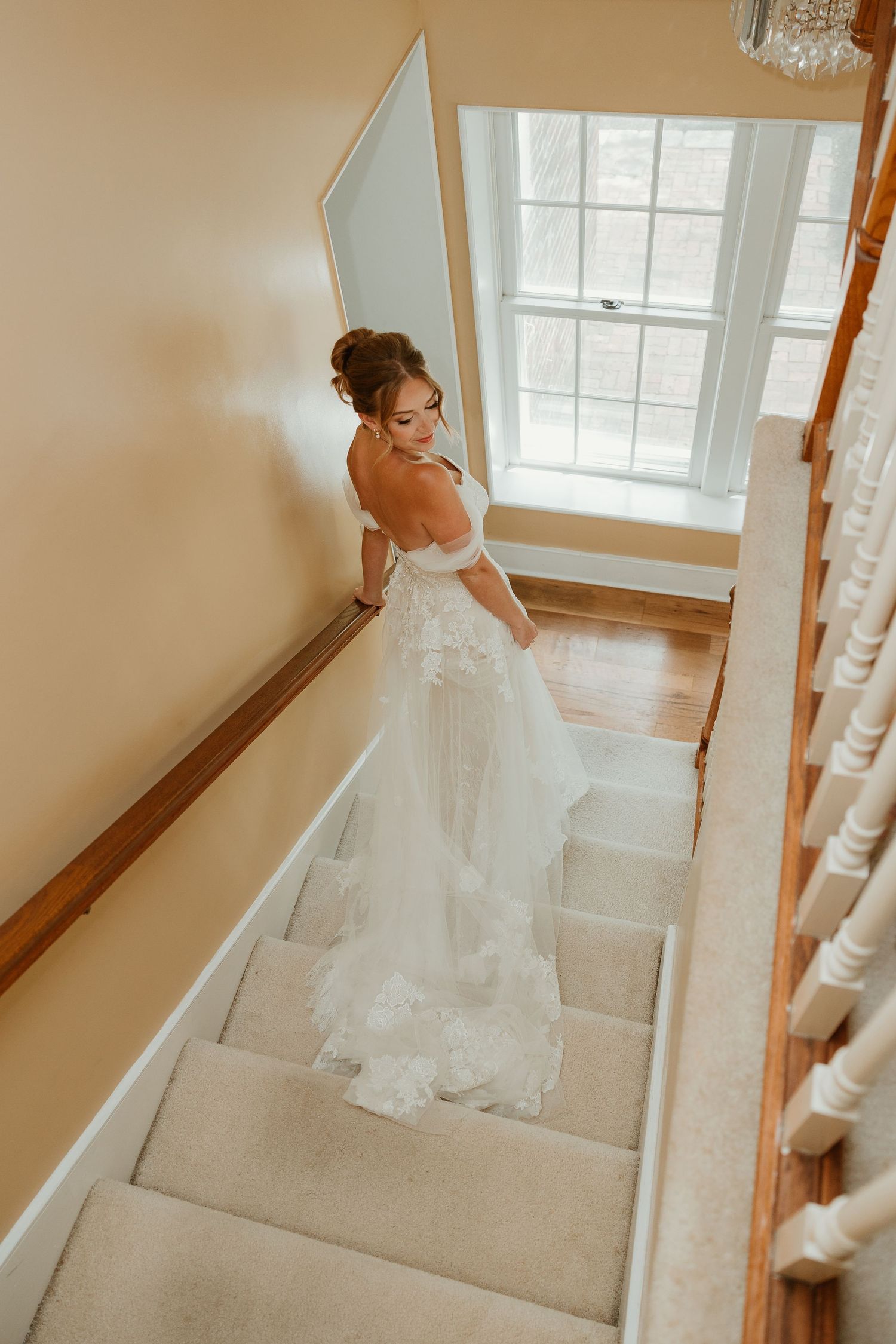 A bride in a flowing white backless gown stands on carpeted stairs near a bright window in warm natural light.