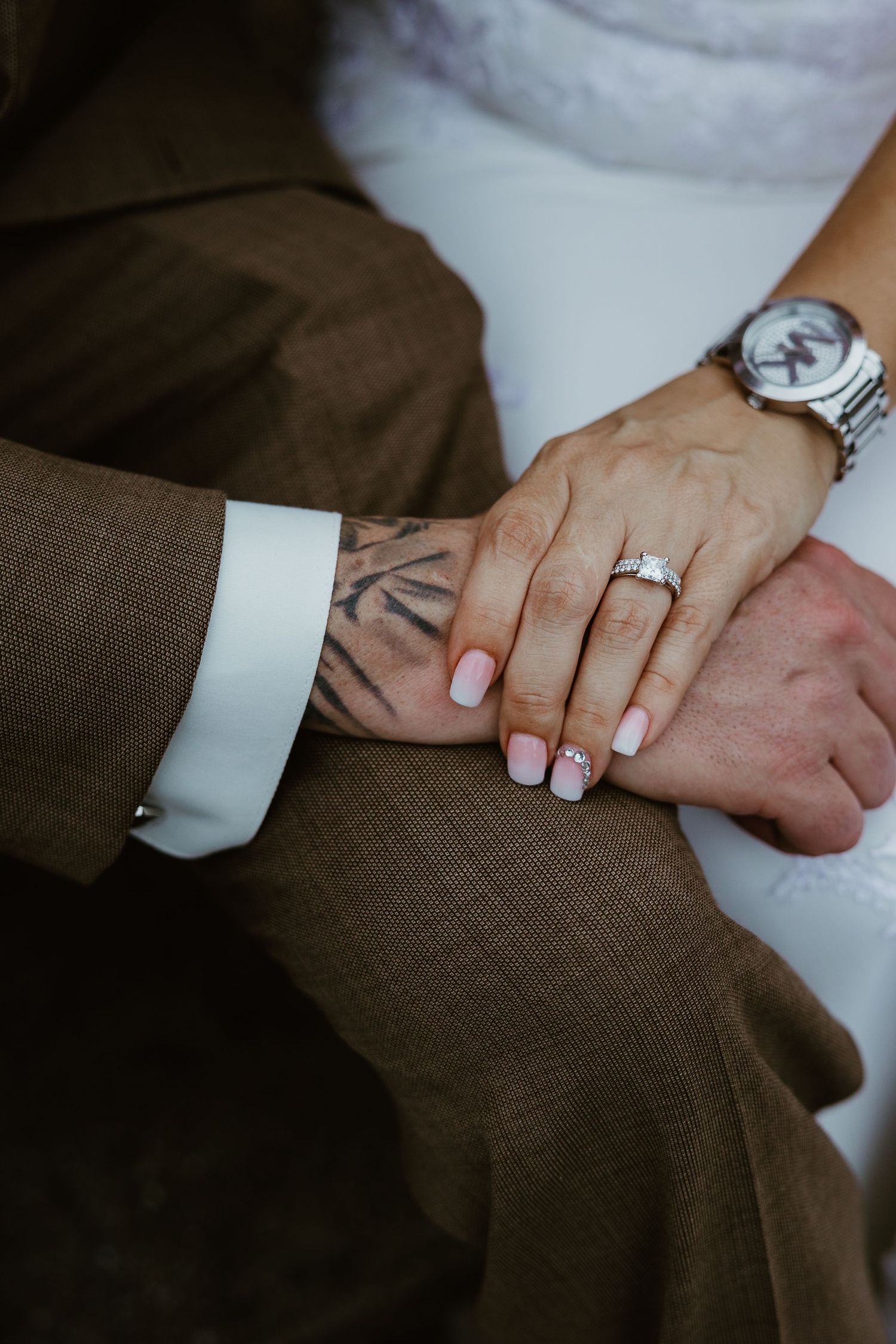 Close-up of hands with engagement ring, silver watch, and tattoo detail against brown suit fabric.
