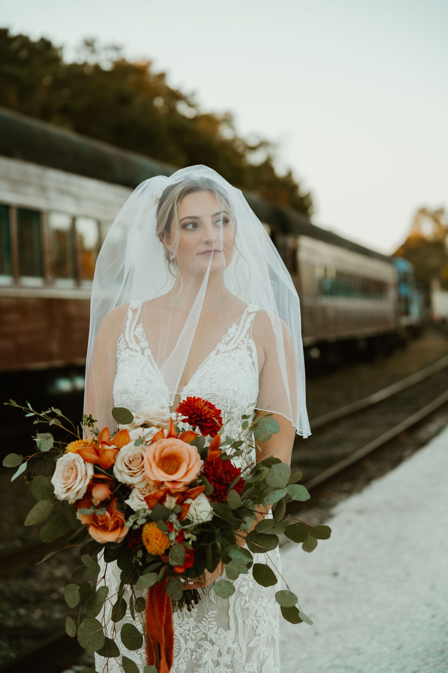 Beautiful bride in lace gown holds vibrant autumn bouquet with red and peach flowers by vintage train.