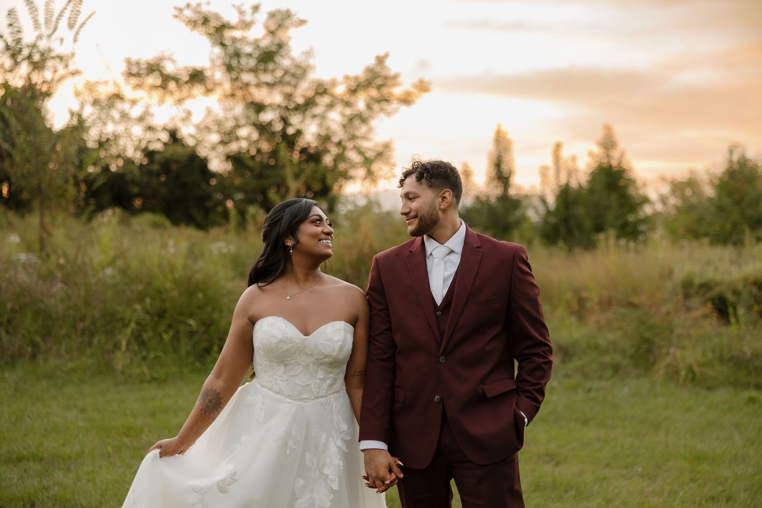 A couple in wedding attire walks together in a golden field during sunset.
