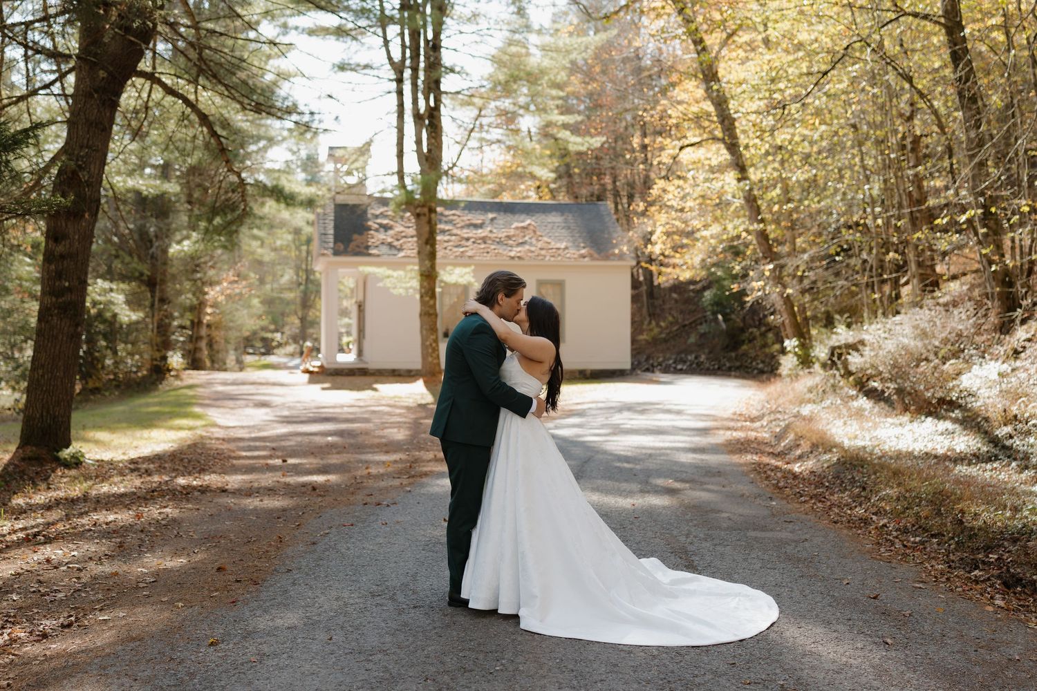 Bride and groom sharing a kiss on a tree-lined path with a charming cottage in the background during fall.