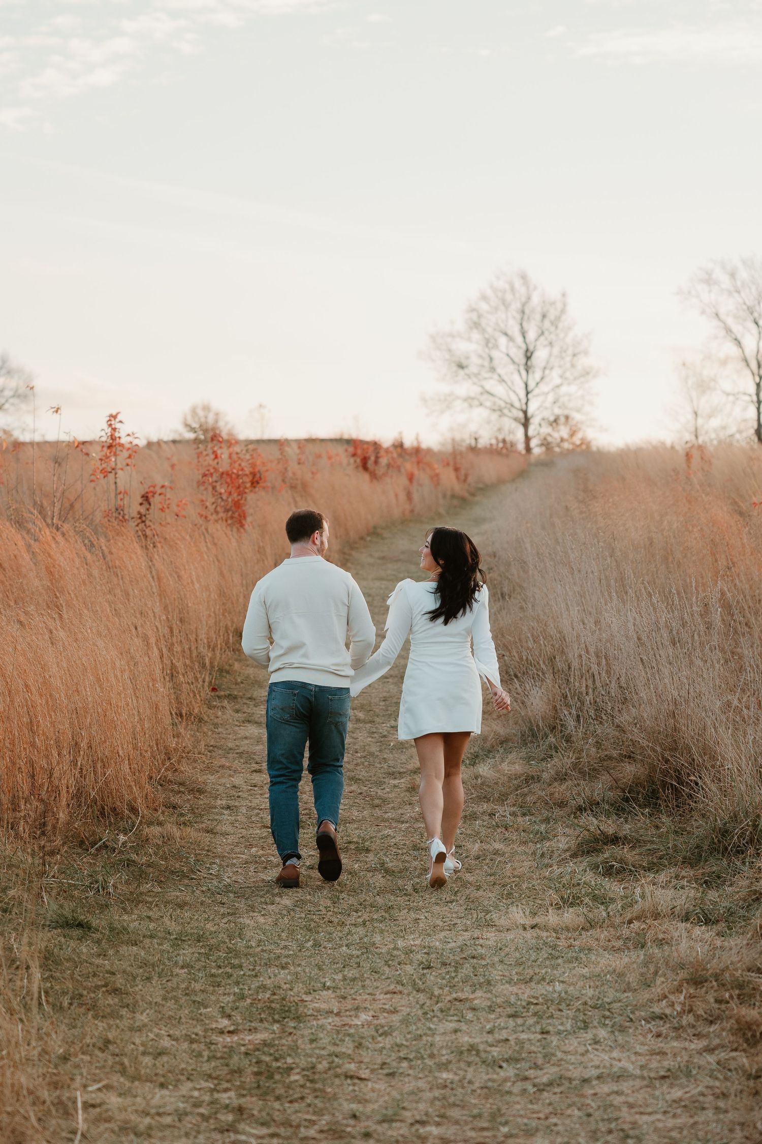A couple walking hand in hand down a dirt path surrounded by tall autumn grass during golden hour.