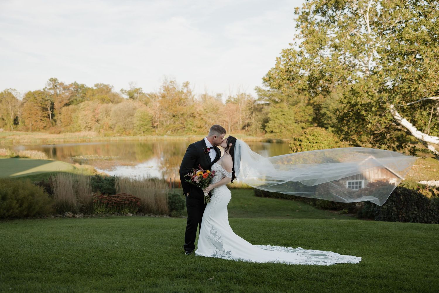 Bride and groom share a romantic kiss on lush green lawn, with veil flowing in wind and serene pond backdrop.