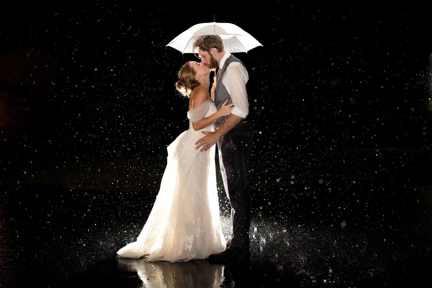 Romantic couple kissing in the rain under a white umbrella, bride in white gown, groom in suit, dark background.