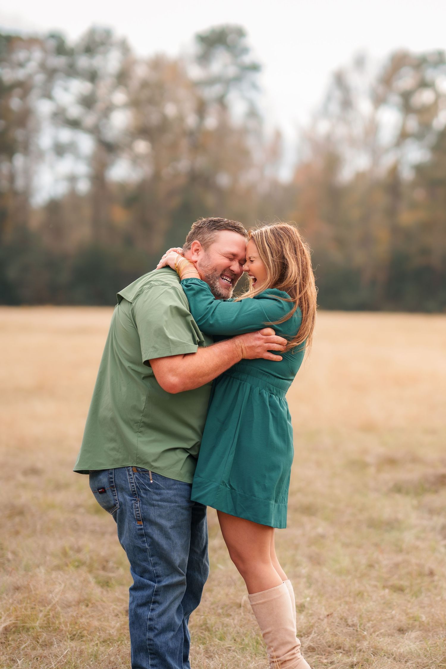 Couple hugging and laughing in an open field with autumn trees, wearing matching green outfits.